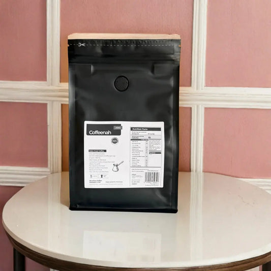 A black coffee bag package with a white label reading 'Coffeemah' stands upright on a round white table against a pink paneled wall.