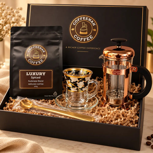 A coffee gift set in a black presentation box, featuring a bag of Coffeenah Luxury Spiced Ground Coffee, a copper-toned French press, a clear glass mug with black and gold patterns, a gold spoon, and shredded paper filler.