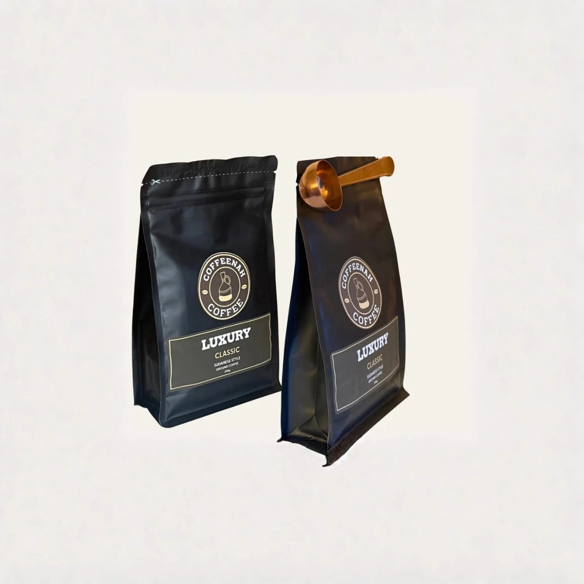 Two black coffee bags labeled 'Coffeera Coffee Luxury' standing upright on a light gray background, with one bag featuring a small metallic gold scoop resting on top.