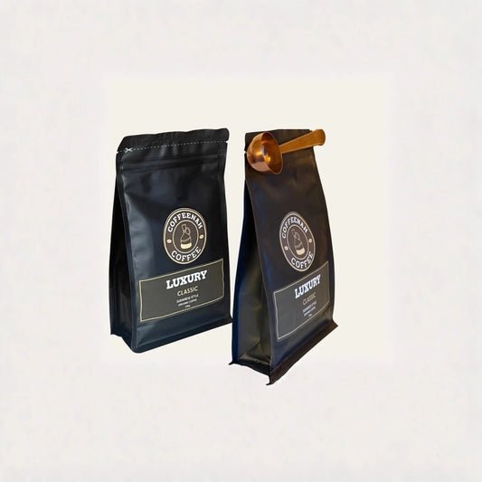 Two black coffee bags labeled 'Coffeera Coffee Luxury' standing upright on a light gray background, with one bag featuring a small metallic gold scoop resting on top.